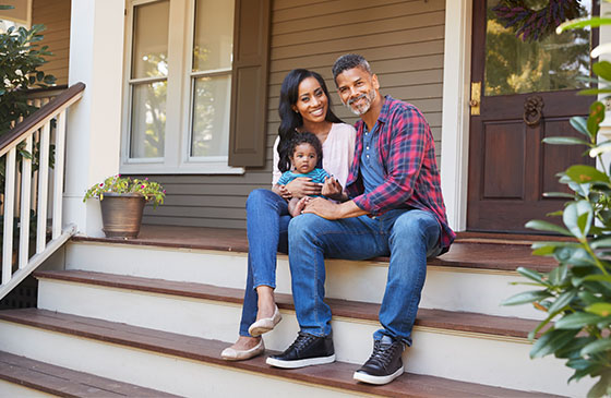 Husband and wife sit with their infant child on the steps of their home.