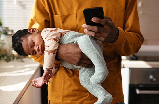Man holding his sleeping baby while doing online banking with his phone. 