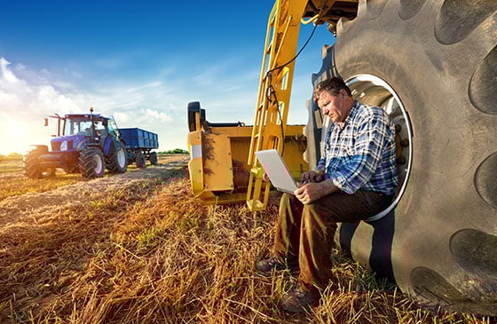 Man looking at his computer while sitting on the tire of farm equipment. 