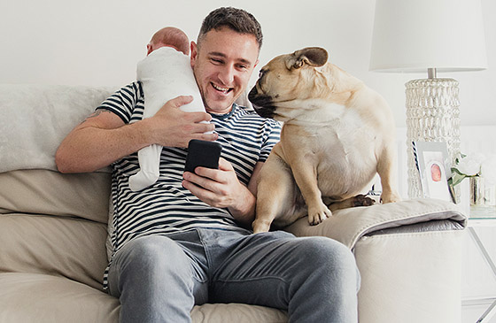 Dad sitting with his newborn child and dog while banking on his phone. 