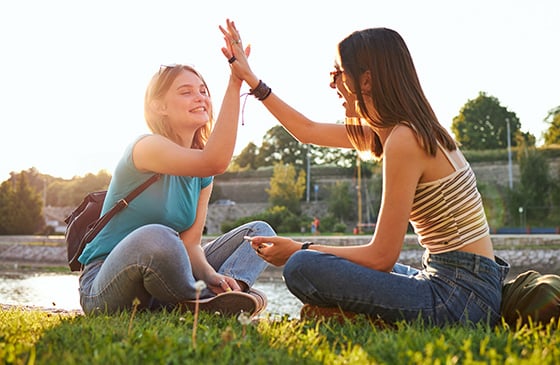 Two girls sitting in a park high fiving. 