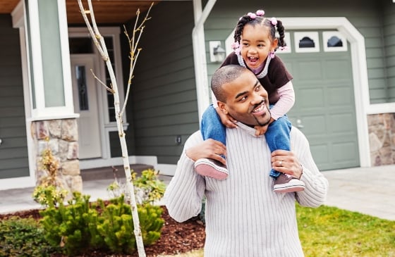 Father with daughter on shoulders in front of a house.