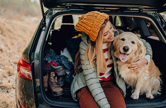 Woman sits with her dog on the trunk of her car.