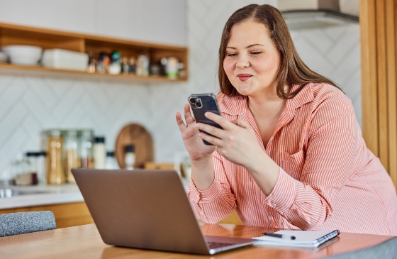 Lady smiling and using her phone with her laptop open in front of her.